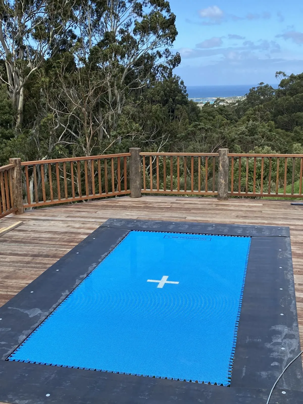 Deck-mounted Mr Trampoline with a bright blue mat set flush into a timber deck, overlooking bushland and the ocean at Apollo Bay