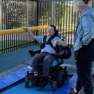 A young person in a power wheelchair on a padded mat beside a trampoline, an adult standing alongside