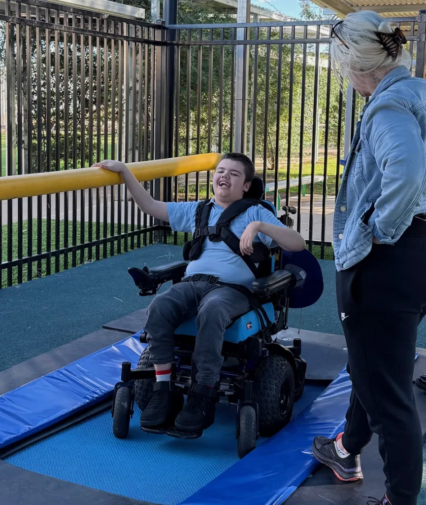A young person in a power wheelchair on a padded mat beside a trampoline, an adult standing alongside