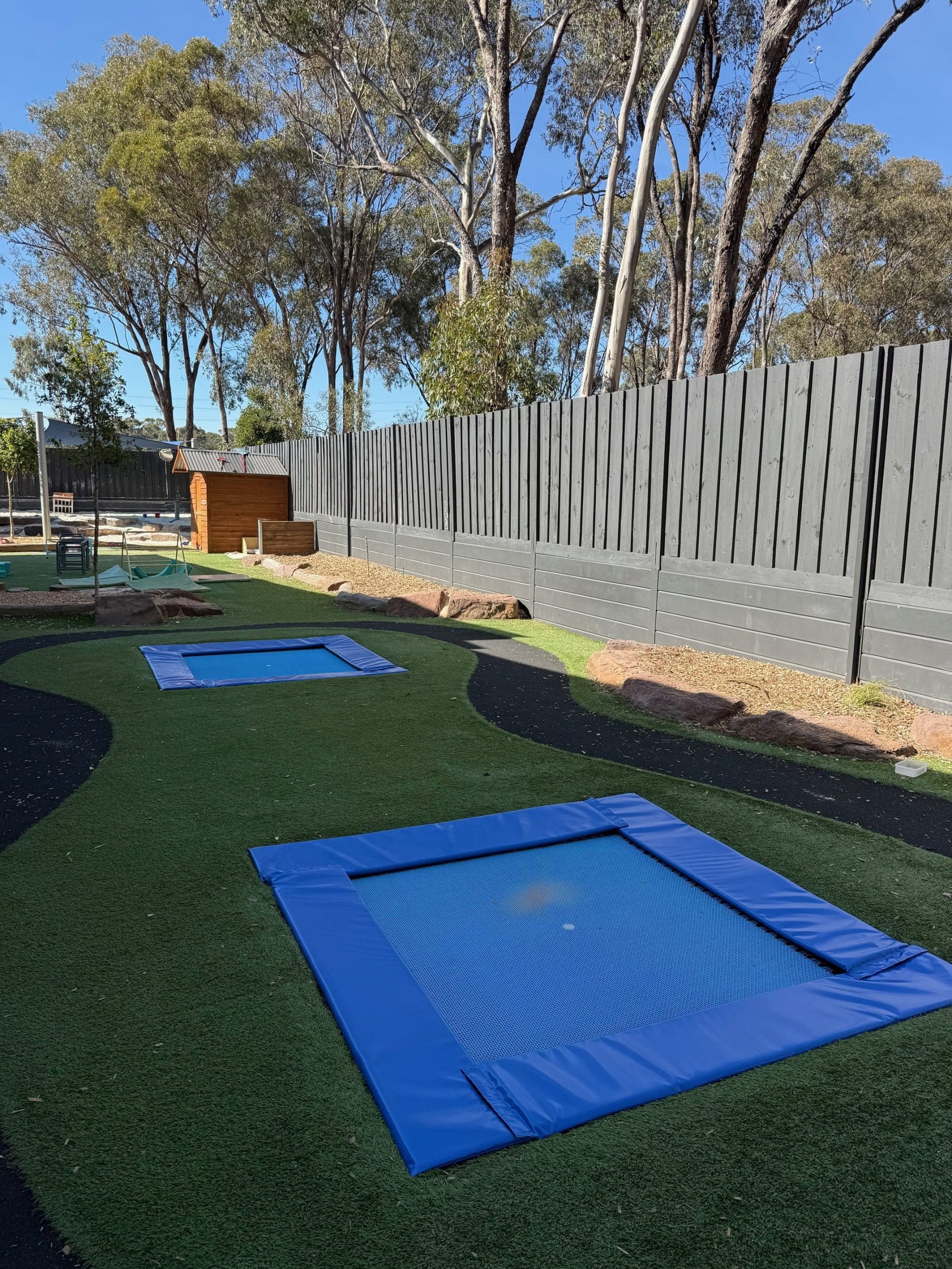 Two blue trampolines on synthetic grass in a fenced yard, one set flush in-ground and one with a padded surround