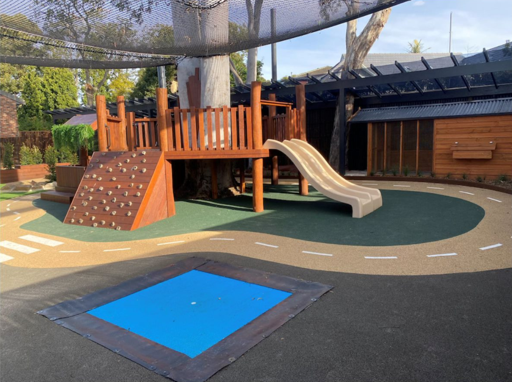 Blue in-ground trampoline set flush into soft-fall rubber in a shaded childcare playground with a timber play structure